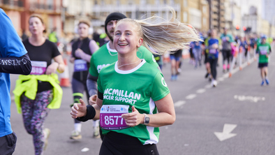 A woman is running in a green Macmillan top. Her blonde hair is pulled back in a pony tail. She appears to be running on a road, and there are other runners next to and behind her.