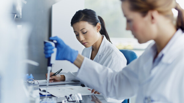 Two female scientists working in a laboratory. One is using a pipette to handle a sample, while the other writes notes on a document. 