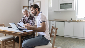 A father and son are talking to one another whilst sat at a dining table. They have a laptop open on the table and some other paperwork around them.