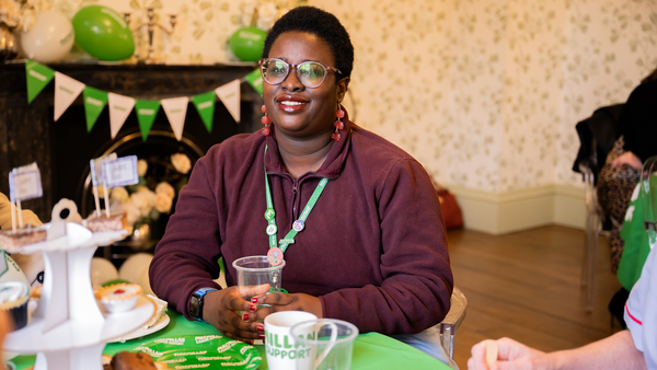 A person with short, dark hair is sitting at a table. They are attending a Coffee Morning and are holding a plastic drink cup in their hands.