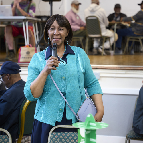 A person is speaking into a microphone at a Coffee Morning. They have short, dark hair and are wearing a bright blue cardigan and dark blue dress. They are standing near a table and speaking to Coffee Morning attendees. 