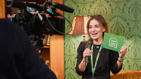 A woman with dark hair is standing in front of a camera. She is holding a microphone and green card which reads 'Open House' 