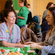 A Macmillan nurse and a Coffee Morning attendee are sat at a table and talking. On the table are different treats and drinks. They appear to be in a conference room filled with other Coffee Morning attendees.