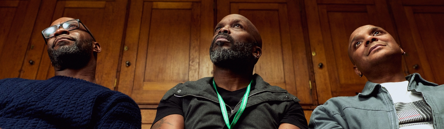 Three people sit side by side in a wood‑panelled indoor setting, conveying a calm, reflective atmosphere within a professional environment. One person is wearing a green Macmillan lanyard.