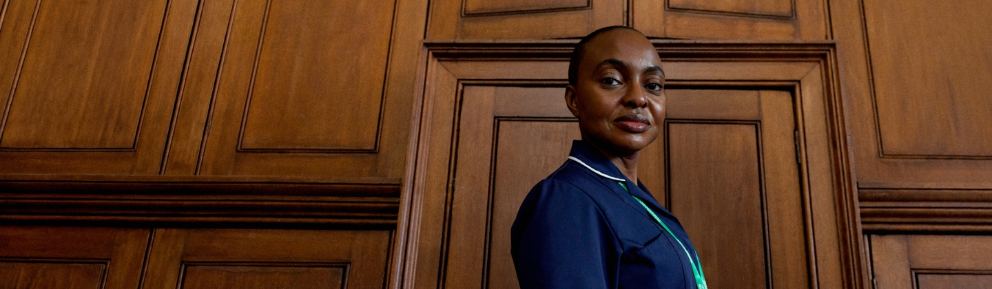 Person wearing a navy uniform and green Macmillan lanyard standing against a wood‑panelled interior.