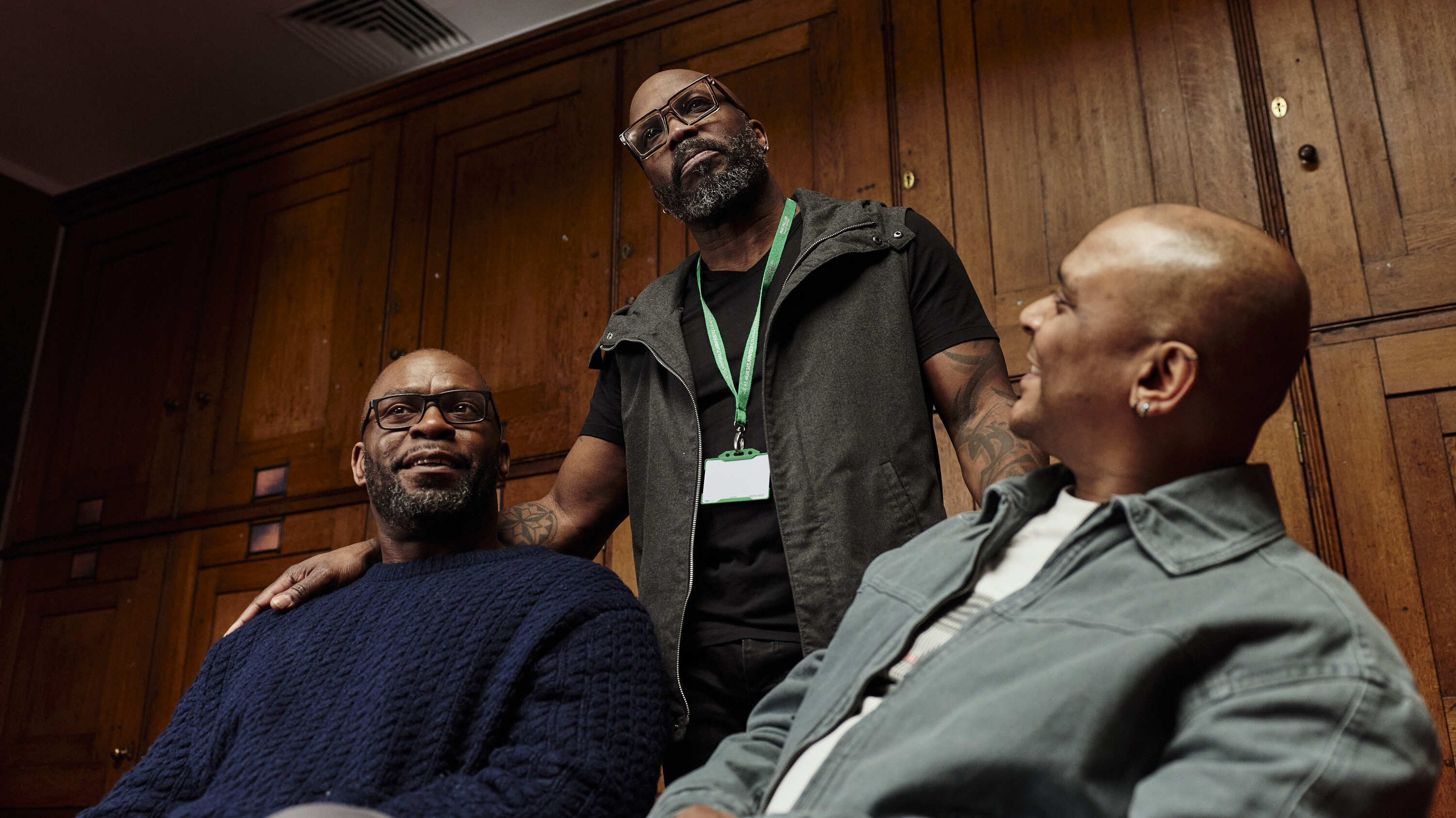 One person stands while others sit in a wood‑panelled room, showing connection and conversation within a support group.