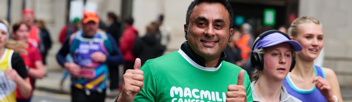 A runner is giving two thumbs up. He is wearing a green Macmillan top and a participation bib pinned to the top. 