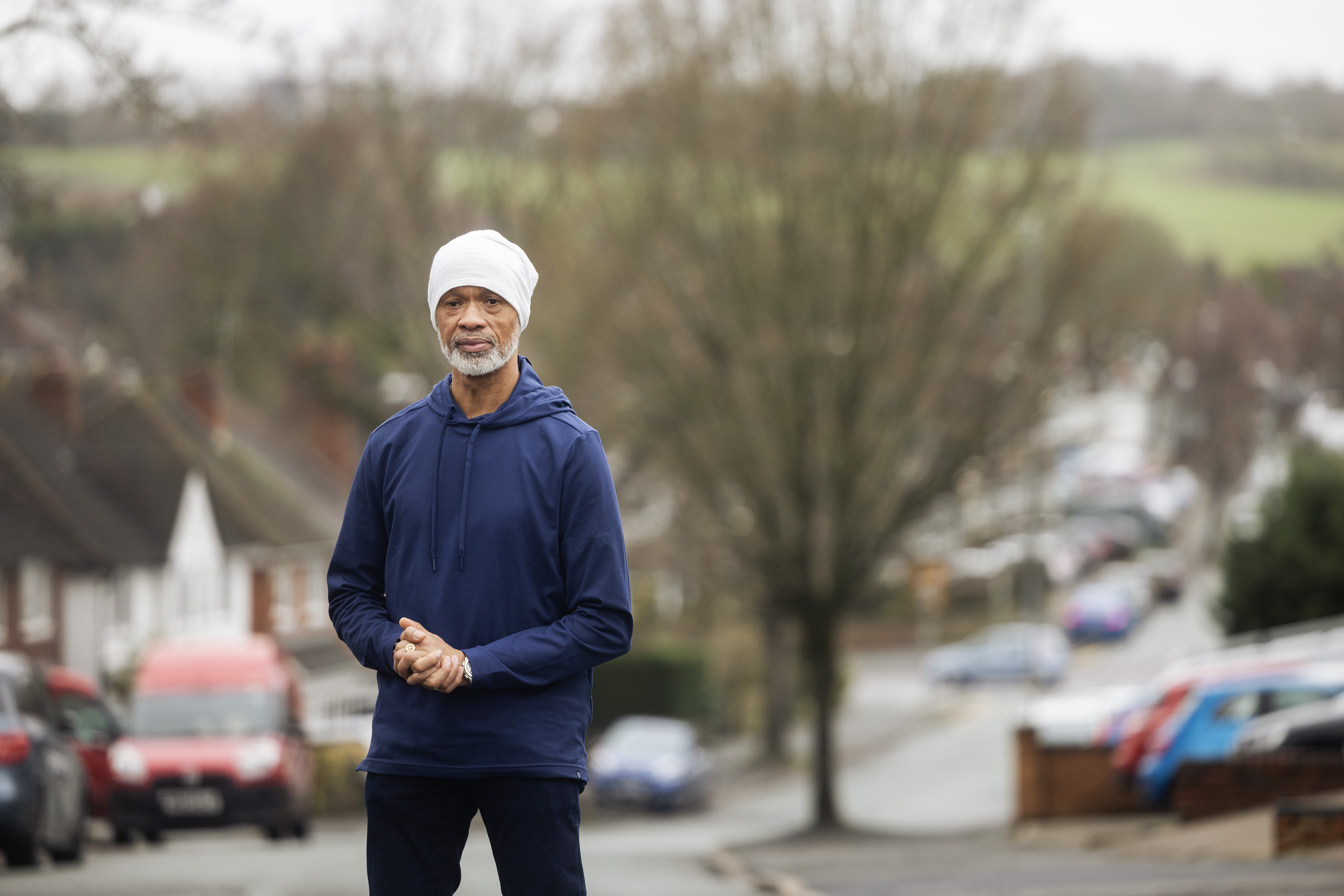 A person stands on a residential street wearing casual clothing, with houses and parked cars in the background.