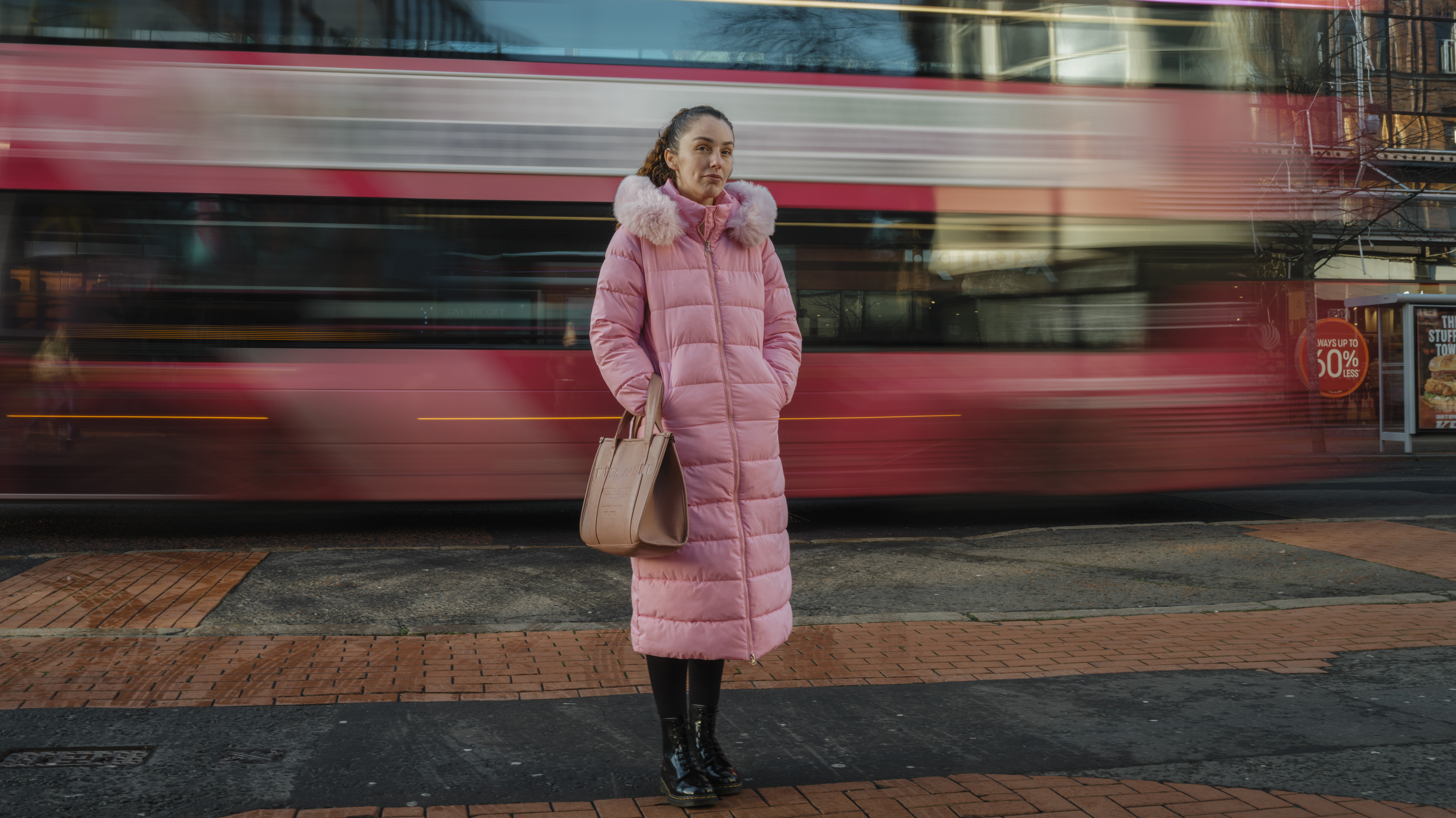 A woman in a long pink coat standing in front of a red bus. The red bus is in motion and blurred.