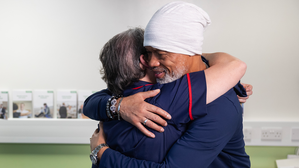 Two people embrace in a clinical meeting room, with posters and leaflets visible on the walls.