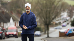 A person stands on a residential street wearing casual clothing, with houses and parked cars in the background.