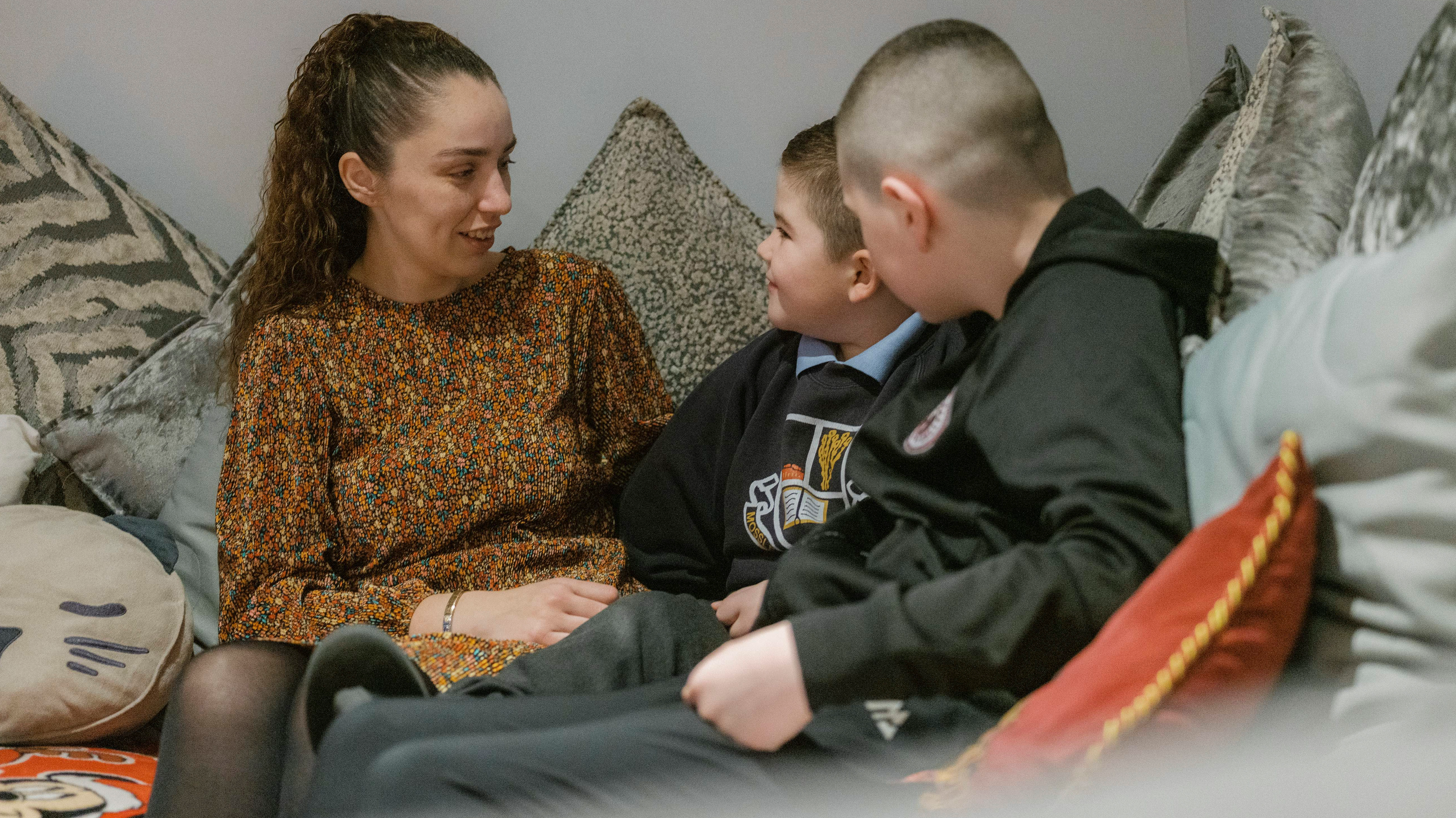 An adult and two children sit together on a sofa in a living room, talking closely and facing one another.