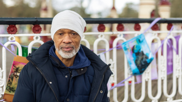 Person standing outdoors by a decorative railing with colourful artwork and ribbons tied along it.