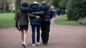 Three people walk together along a park path with their arms around each other, supported by a green, tree-lined setting.