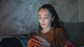 A person sits on a sofa at home, holding a smartphone and looking at the screen.