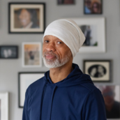 A person wearing a head covering stands indoors in front of a wall filled with framed photographs.