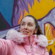 A person in a pink coat holds up a smartphone to take a photo in front of a colourful street mural.


