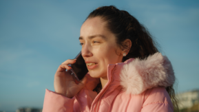 A person wearing a pink coat talks on a mobile phone outdoors near water under a blue sky.