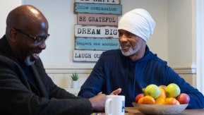Two people sit at a kitchen table talking, with a bowl of fruit and mugs on the table and a sign with positive messages on the wall behind them.
