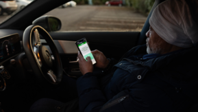 A person sits in the driver’s seat of a parked car, holding a smartphone and looking at the screen.   