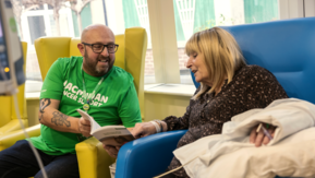 A Macmillan Cancer Support volunteer sits beside someone in a hospital chair, looking at a booklet together.