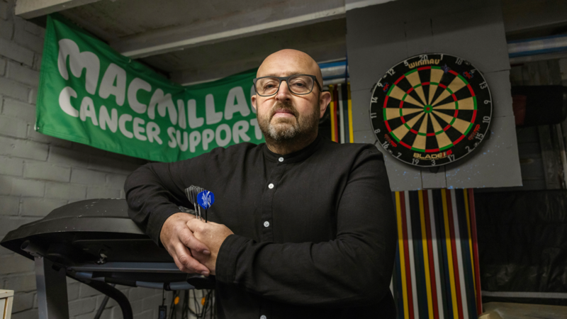 A person stands indoors with arms folded, with a Macmillan Cancer Support banner and dartboard behind them.


