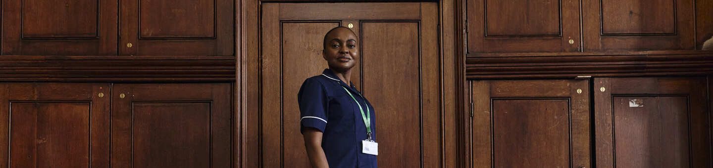 A nurse stands in profile against a wood‑panelled interior, wearing a uniform and lanyard in a quiet indoor space.