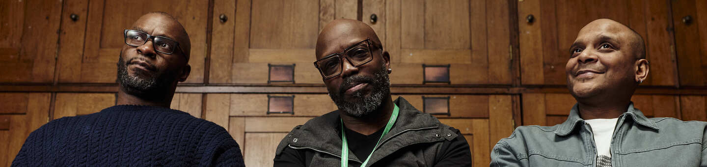 Three people sit side by side in a wood‑panelled room. The person in the middle is wearing a green Macmillan lanyard.