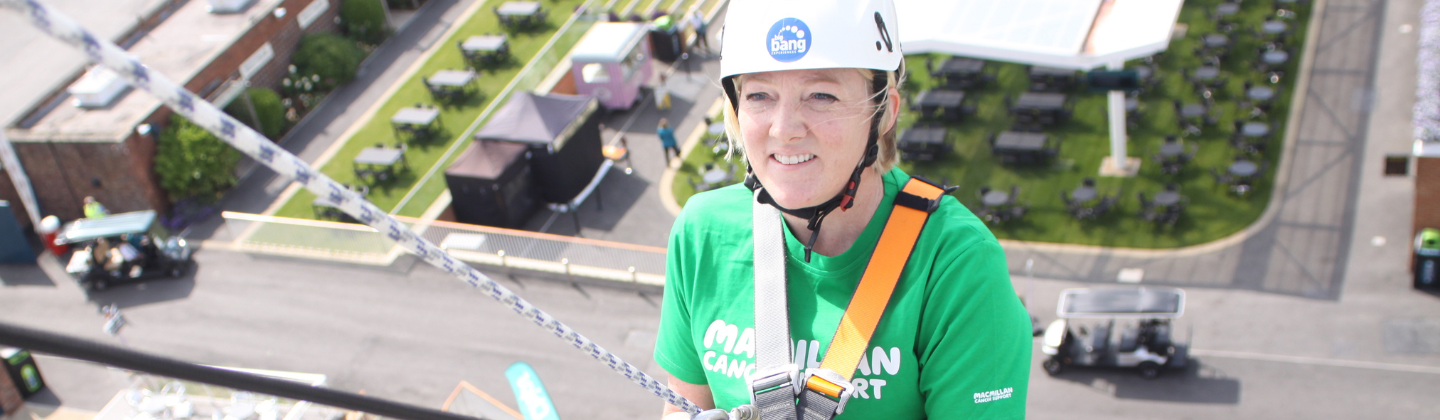 A person abseiling. They are wearing a helmet and a green Macmillan branded top. She is in a harness. 