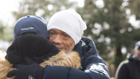 Two people embrace outside. They are wearing puffer coats, hats and gloves indicating a cold but bright day.