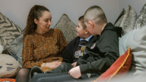 An adult and two children sit together on a sofa in a living room, talking closely and facing one another.