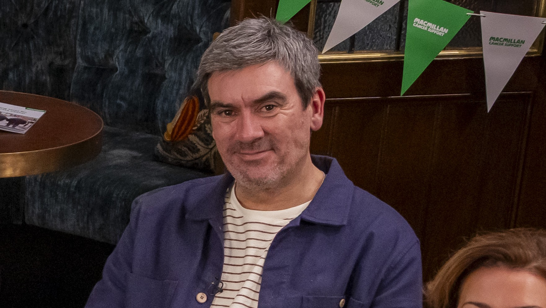 A smiling man seated in a pub-style setting, with tables, seating and Macmillan-branded bunting behind him.
