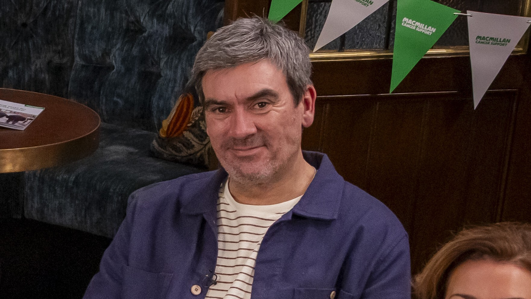 A smiling man seated in a pub-style setting, with tables, seating and Macmillan-branded bunting behind him.