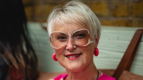 Penny has short hair and is wearing white rimmed glasses. She has on a pink and white patterned top and pink earrings. She appears to be sitting somewhere indoors.
