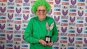 John is standing in front of a Pride of Britain backdrop. He is wearing a bright green curly wig and a green long sleeved top. In his hands he is holding a trophy. 