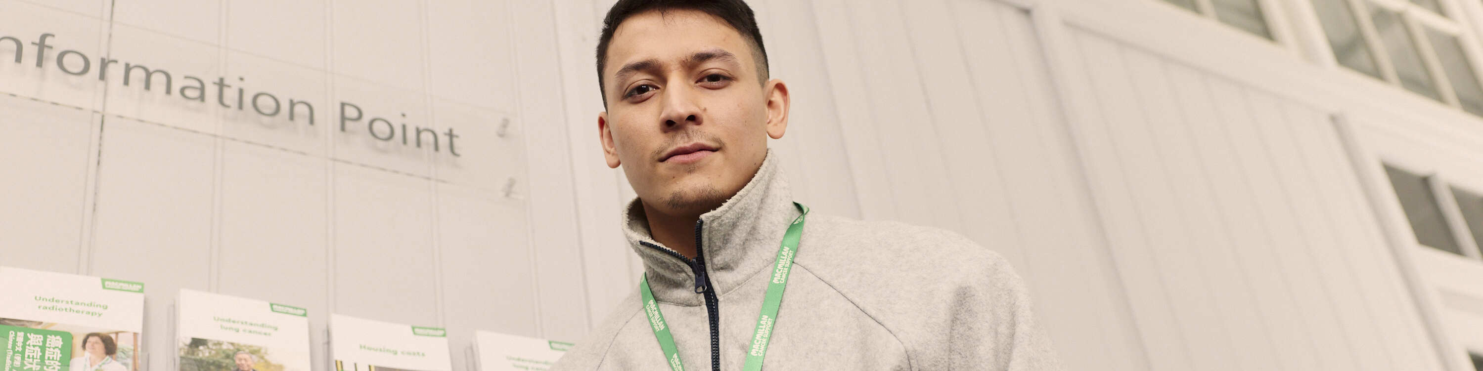 A person in a wheelchair with a person beside them wearing a green lanyard. They are in an indoor information point, with leaflets displayed on the wall.