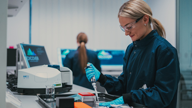 A scientist is using a transfer pipette in a laboratory. They are wearing a dark blue lab coat and safety goggles. Behind them is other lab equipment like computers.