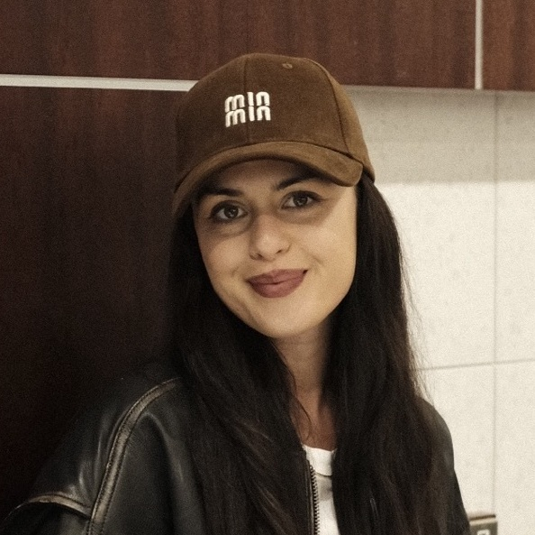 Woman sitting on a wooden bench indoors, wearing a cap and jacket, holding a cup.