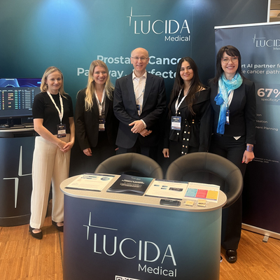 Dr Anthony Rix is standing in the middle of a row of people. He has short hair and glasses and is wearing a shirt and suit. In front of him is a stand for Lucida Medical.