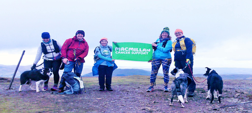 Becky is standing with her friends at completing a hike. She is holding up a green Macmillan sign with a friend. They are all wearing hiking gear and a few are partnered with their pet dogs.