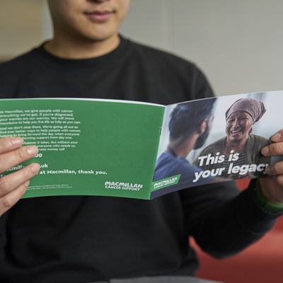 A person is holding open a Macmillan Gifts in Wills booklet. They are wearing a dark top. The booklets cover and the back of the booklet can be seen.