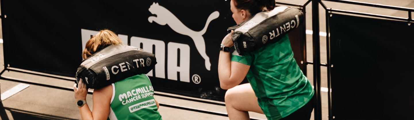 Two participants are taking part in a HYROX. They are carrying sand bags on their backs and are doing lunges. Both are wearing a green Macmillan branded top.