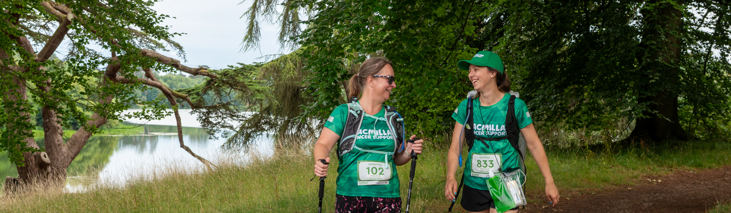 Two hikers are walking along a dirt path near a lake. They are both wearing green Macmillan branded tops.