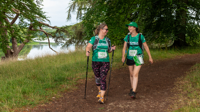Two hikers are walking along a dirt path near a lake. They are both wearing green Macmillan branded tops.