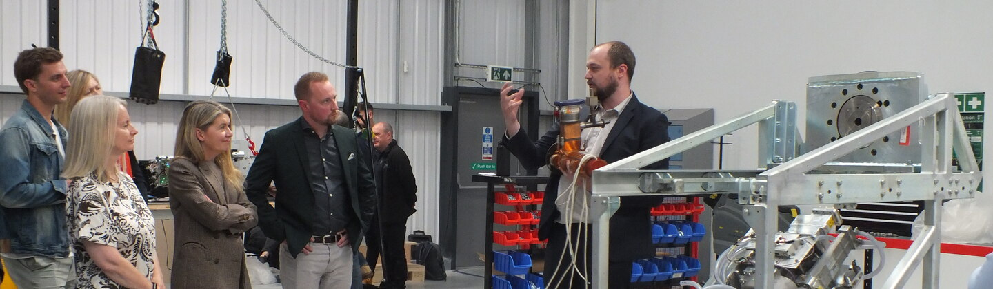A group of people are listening to a person presenting. They appear to be in a warehouse. The person presenting is standing next to a large machine. 