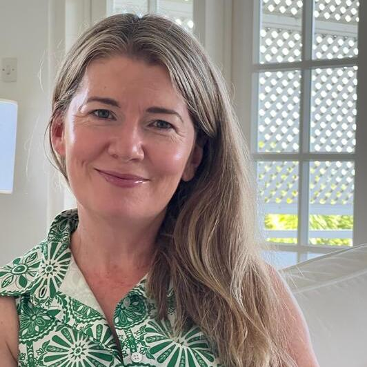 Michelle is sitting on a white sofa and is holding a mug in her hands. She has long hair and is wearing a green and white patterned dress. She appears to be indoors.