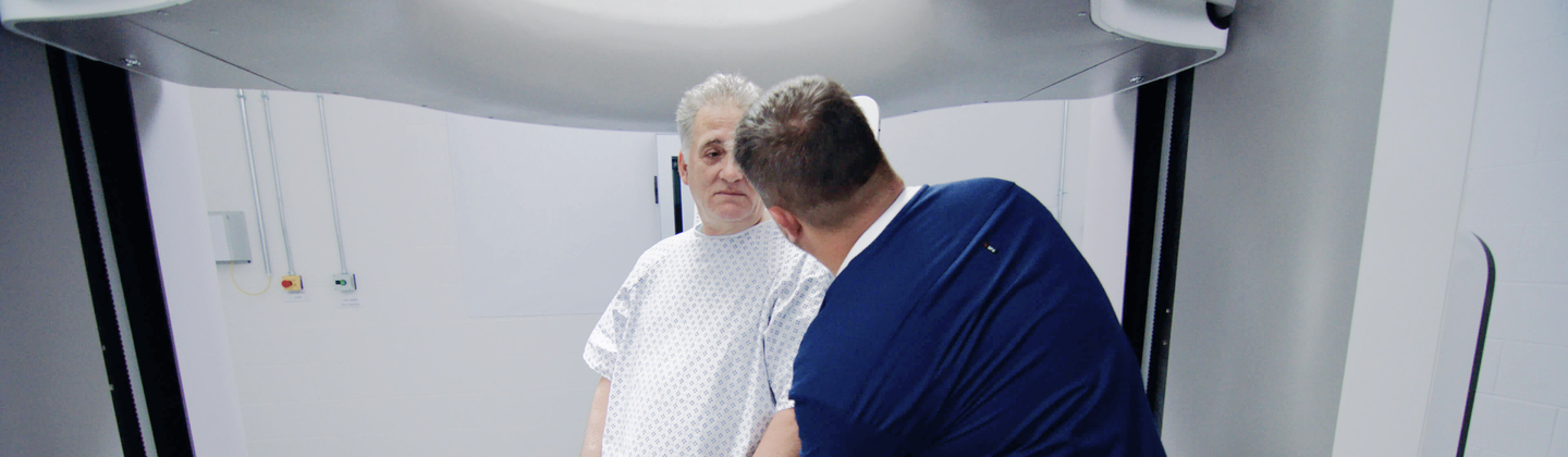 A person is helping a patient who is waiting inside a large scanning machine. The patient is wearing a hospital gown and the person helping is wearing blue scrubs.