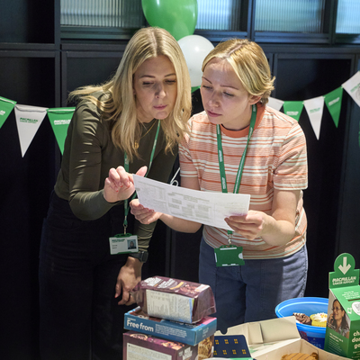 Two people are concentrating on a Coffee Morning quiz sheet. They are standing behind a table full of delicious treats for the event including cakes, muffins, doughnuts and more. 