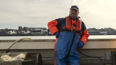 A fisherman leaning against the side of his boat in fishing gear.