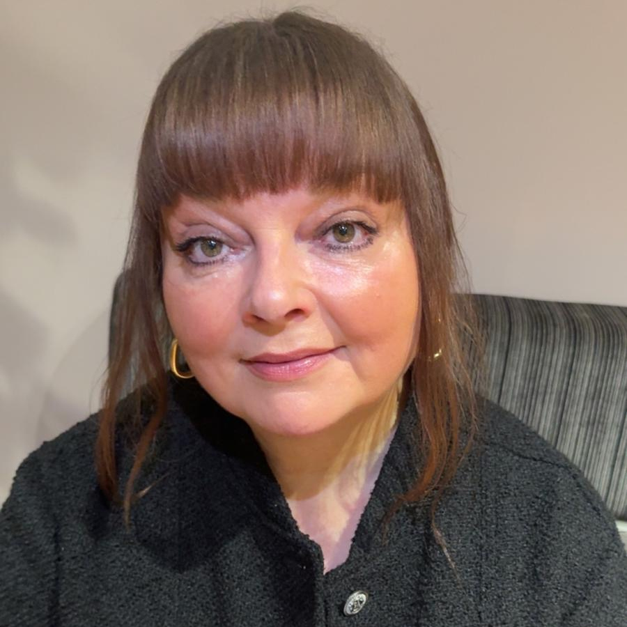 Close-up portrait of a person with straight fringe hairstyle sitting indoors, wearing a dark textured jacket and hoop earrings.